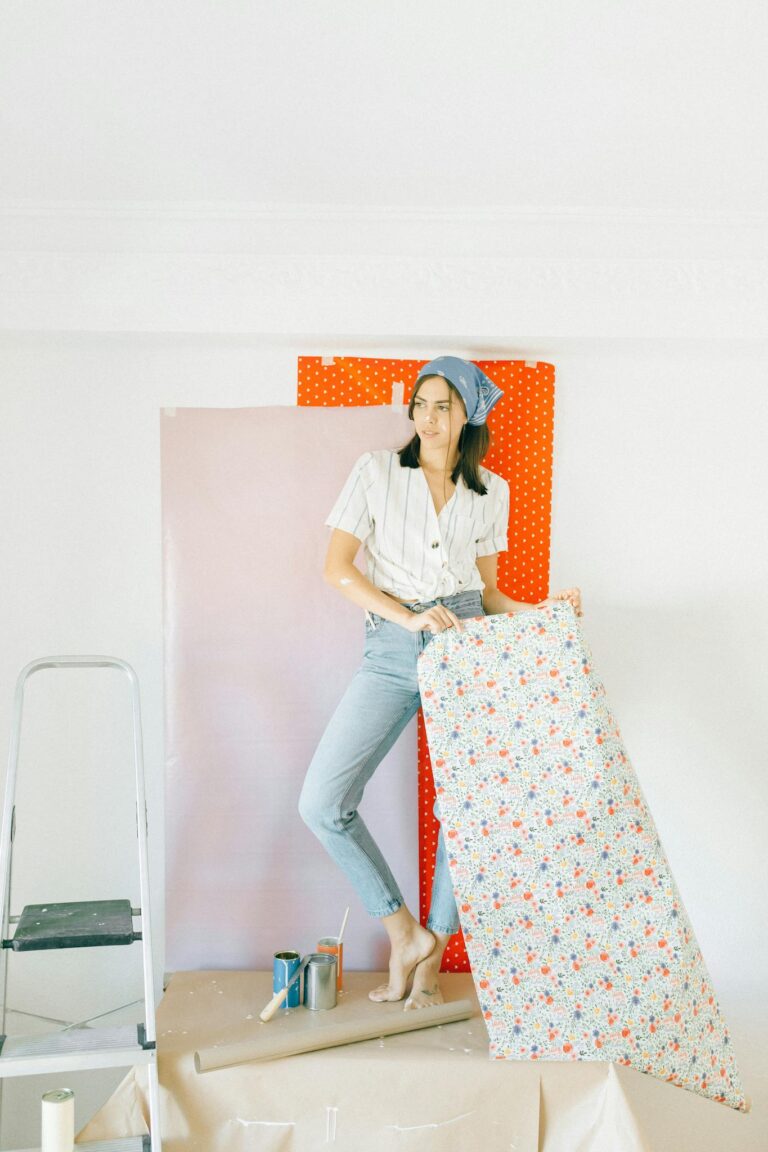 Young woman decorating room with floral wallpaper, standing next to ladder.